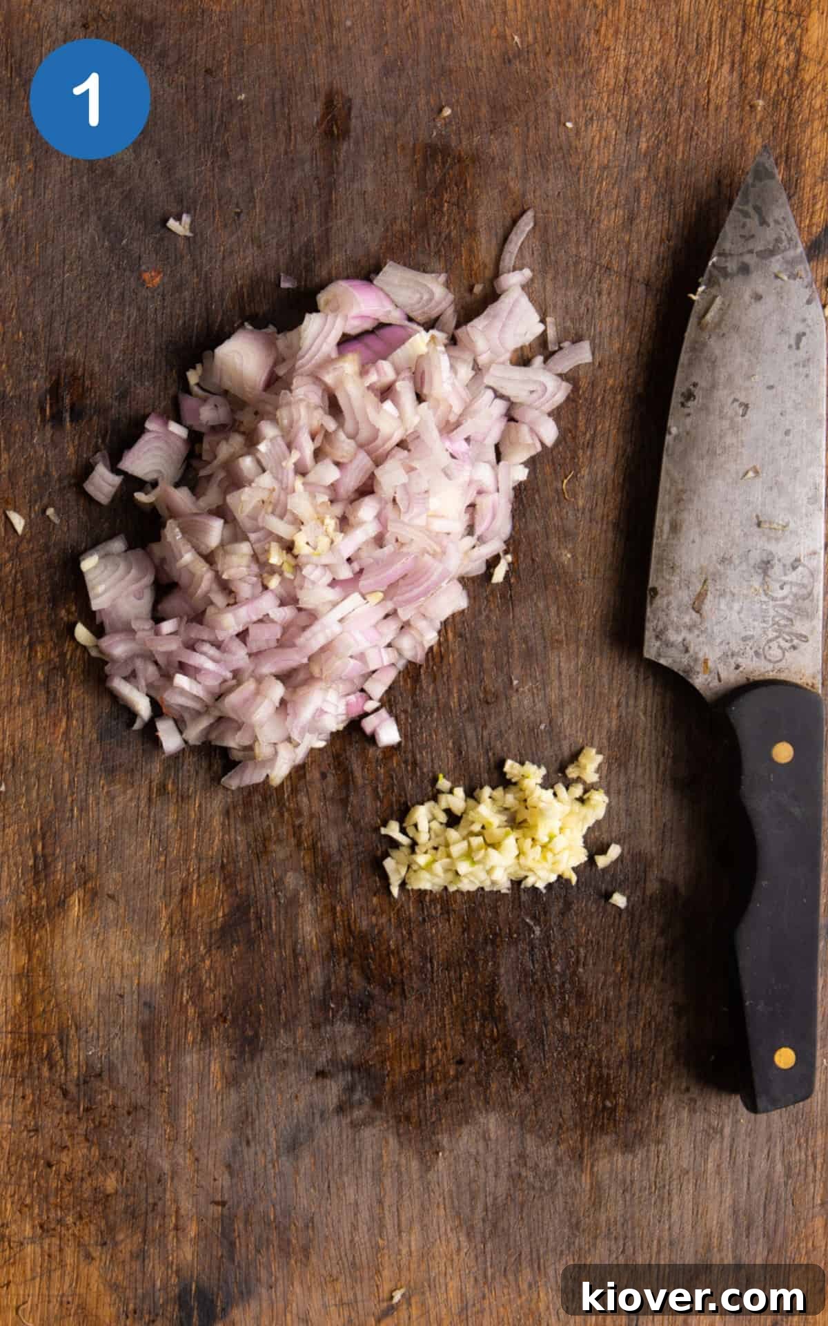 Finely diced shallot and garlic cloves ready on a wooden chopping board next to a knife.