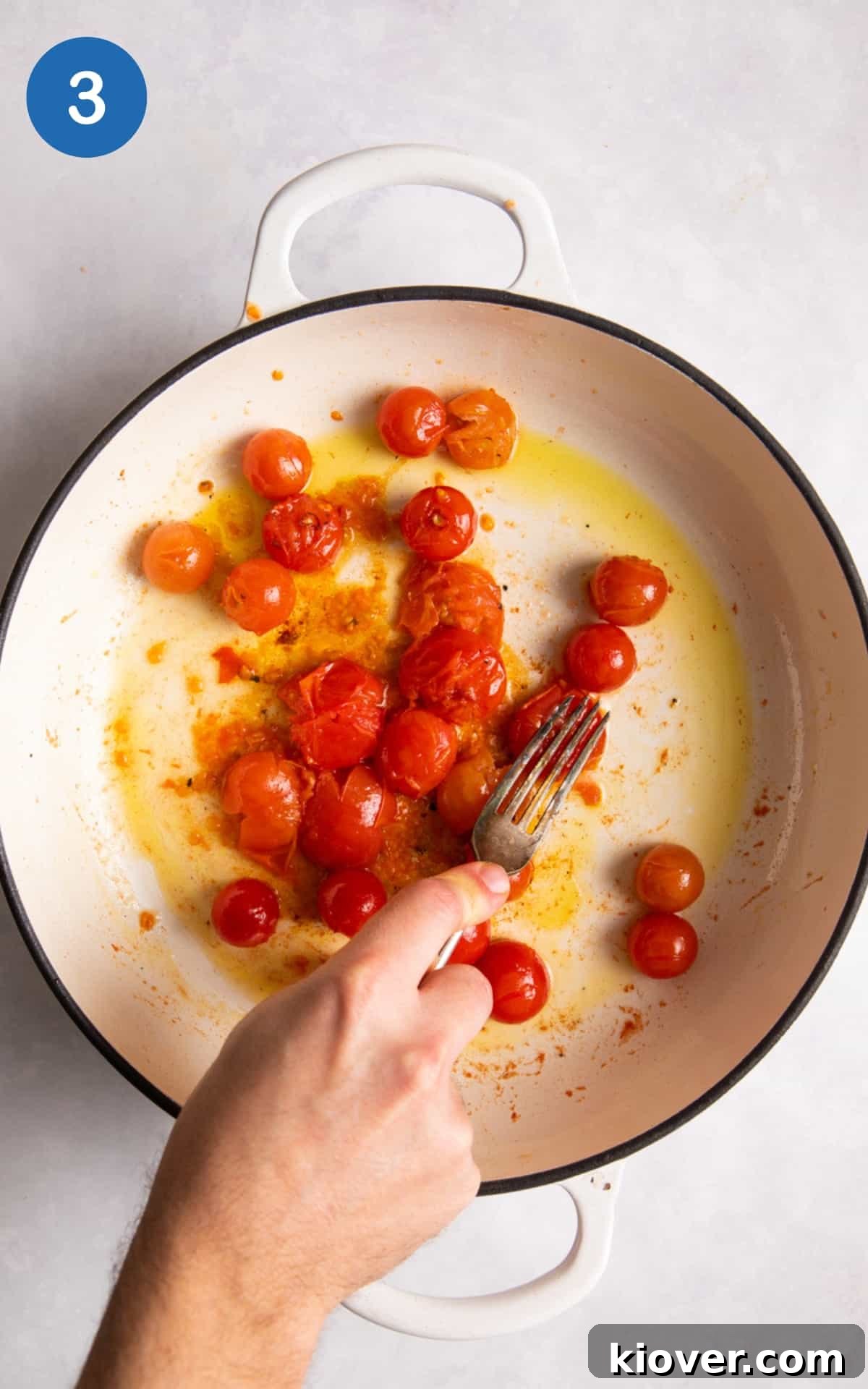 Cherry tomatoes being gently squashed with a fork in a large white pan, releasing their juices to create a sauce base.