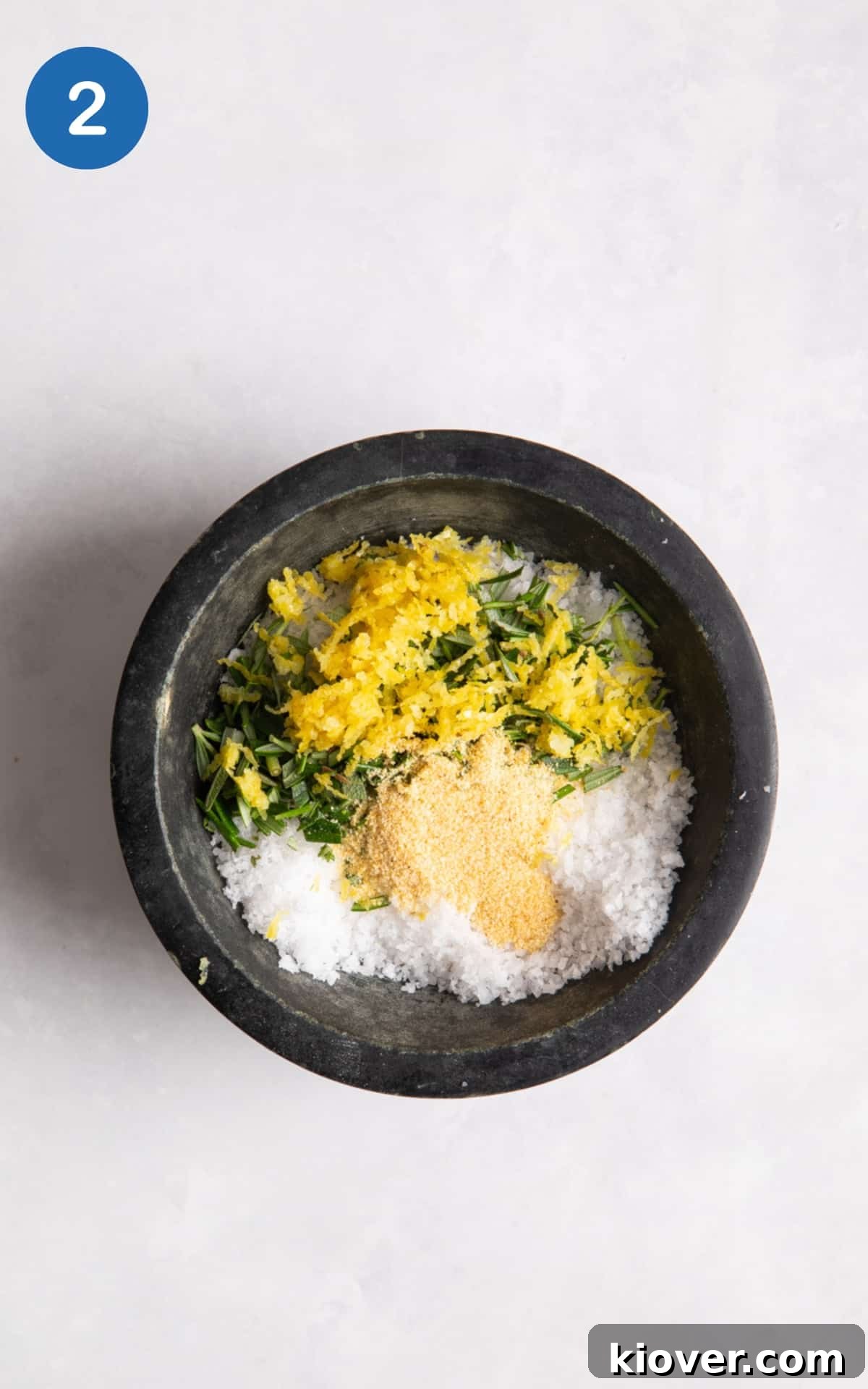 Various ingredients, including chopped rosemary, lemon zest, and garlic granules, being added to a small black pot of flaky sea salt, all placed on a white surface, illustrating the mixing process.