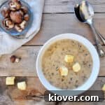 Overhead shot of cream of mushroom soup with croutons on top and fresh mushrooms on the side, presented on aged wood.