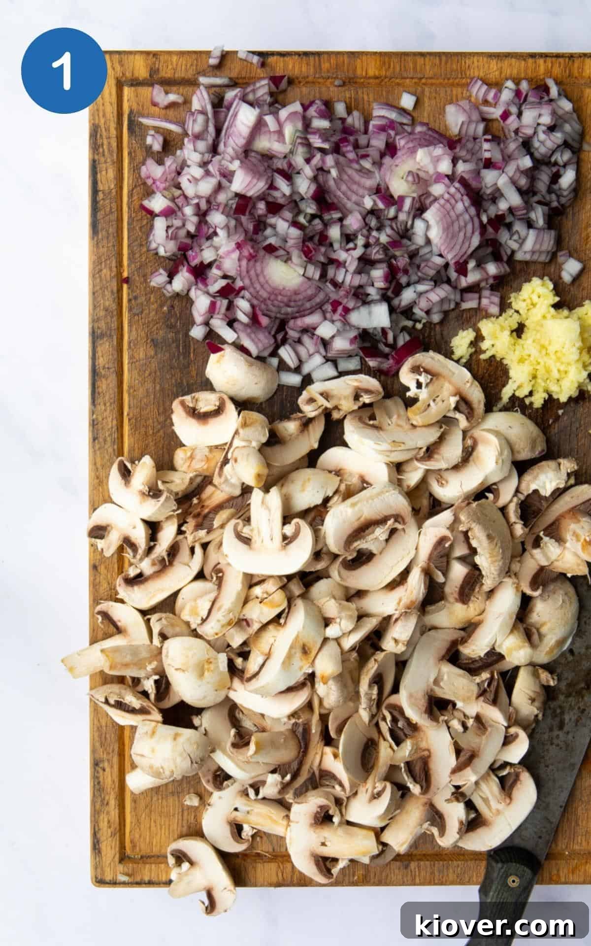 Preparing ingredients: Sliced onion, freshly crushed garlic, and roughly chopped mushrooms are neatly arranged on a wooden chopping board, alongside a sharp kitchen knife. The chopping board rests on a white marble background.