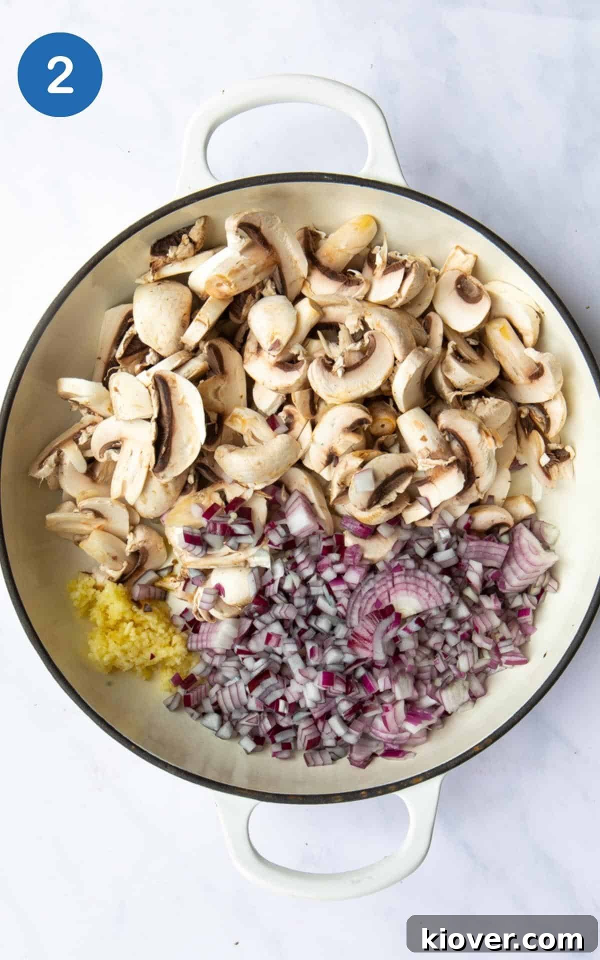 Sautéing the base: Mushrooms, garlic, and onions are actively cooking in a large, shallow frying pan. The pan is positioned on a clean white marble background, indicating the early stages of the stroganoff preparation.