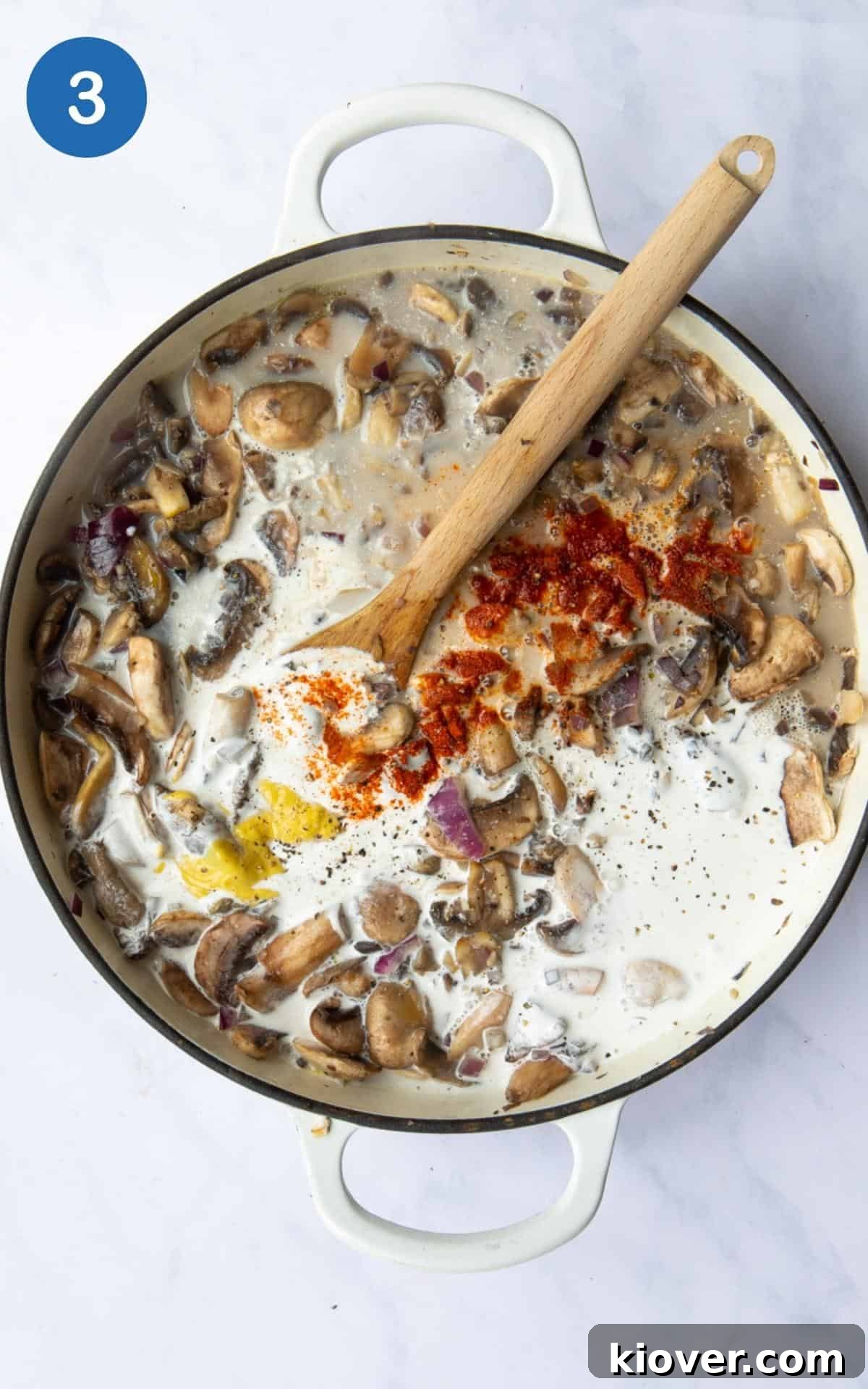 Creamy sauce development: Mushrooms, onion, garlic, coconut milk, paprika, and Dijon mustard are combined in a pan, actively simmering. A wooden spoon rests in the pan, suggesting it's being stirred to combine the rich ingredients. The pan is set against a white marble background.