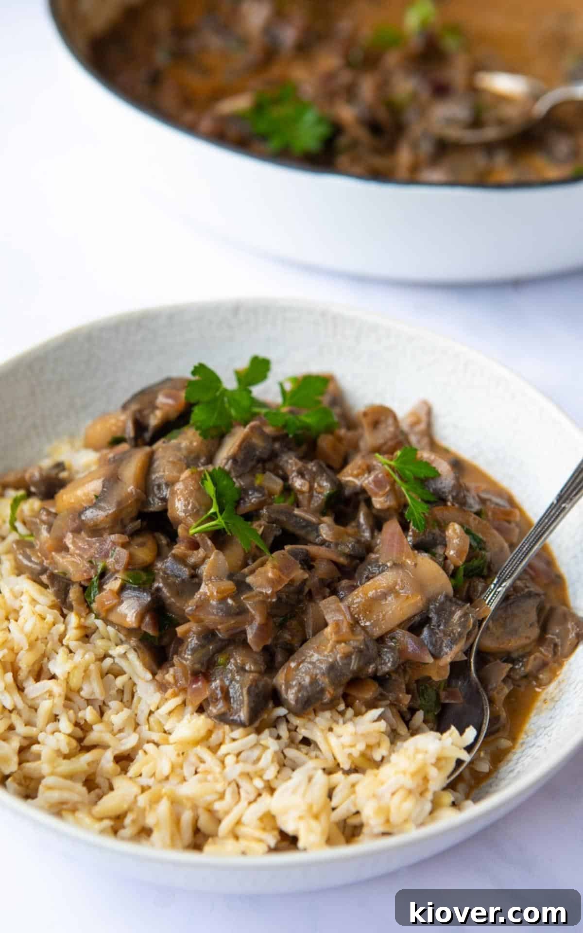 Creamy Vegan Mushroom Stroganoff elegantly served in a white bowl, accompanied by a portion of wholesome brown rice. The bowl is meticulously presented on a sophisticated white marble background, highlighting the dish's inviting appeal.