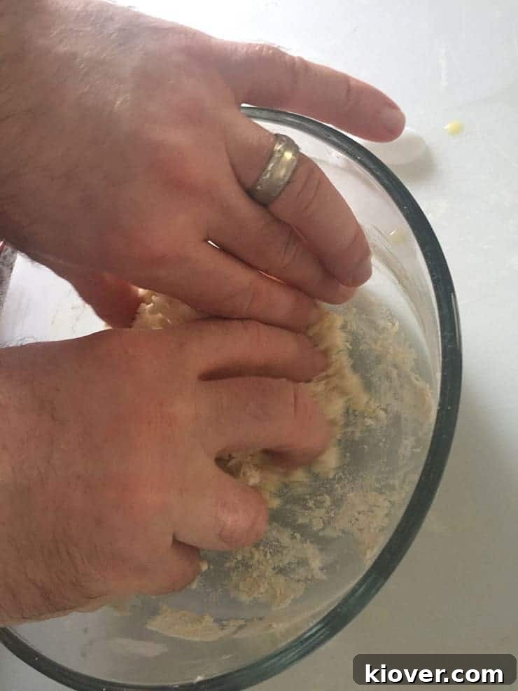 A skilled hand is seen kneading dough vigorously within a clear glass bowl, showcasing the initial stages of preparing homemade noodles.