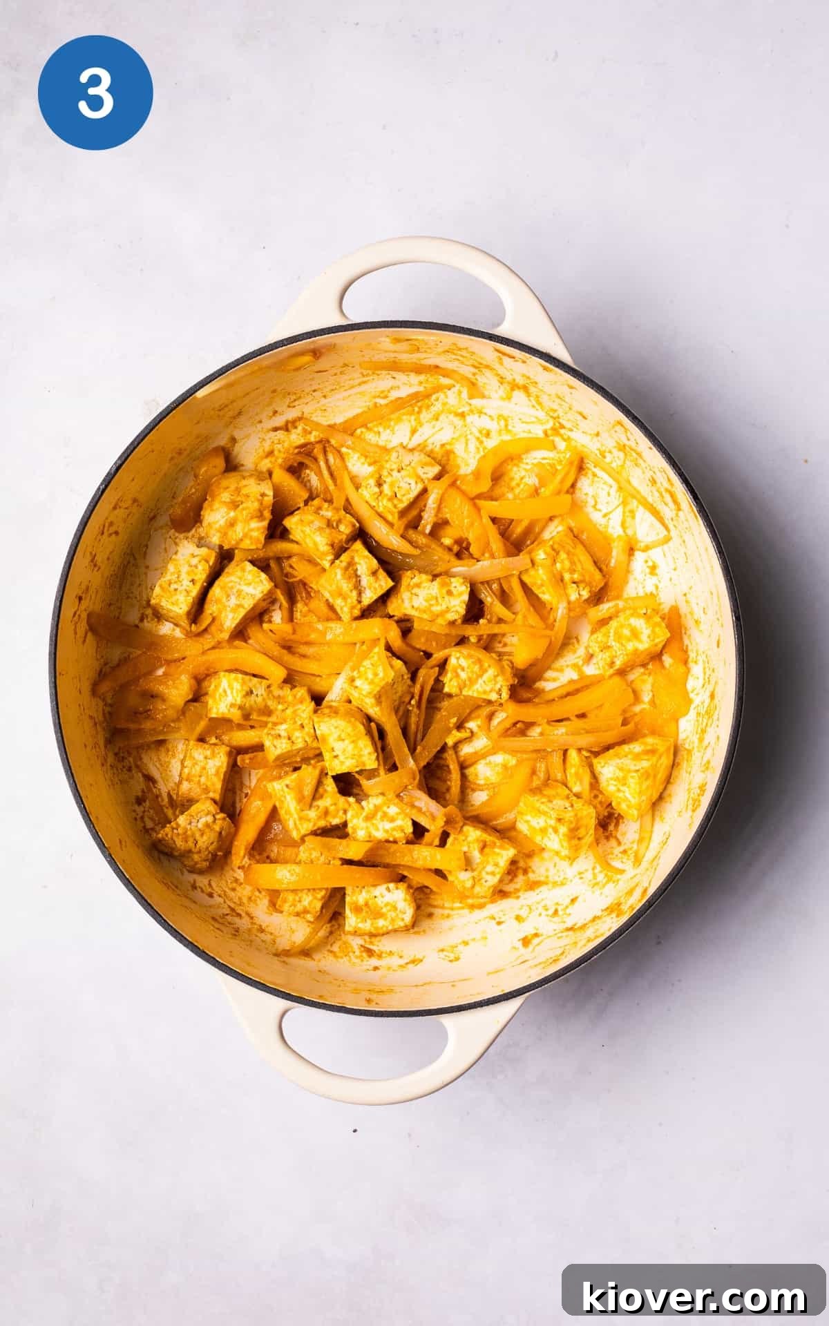 Cubed tofu being mixed into the sautéed shallots, bell peppers, and laksa paste in a pot, ensuring even coating and absorption of spices.