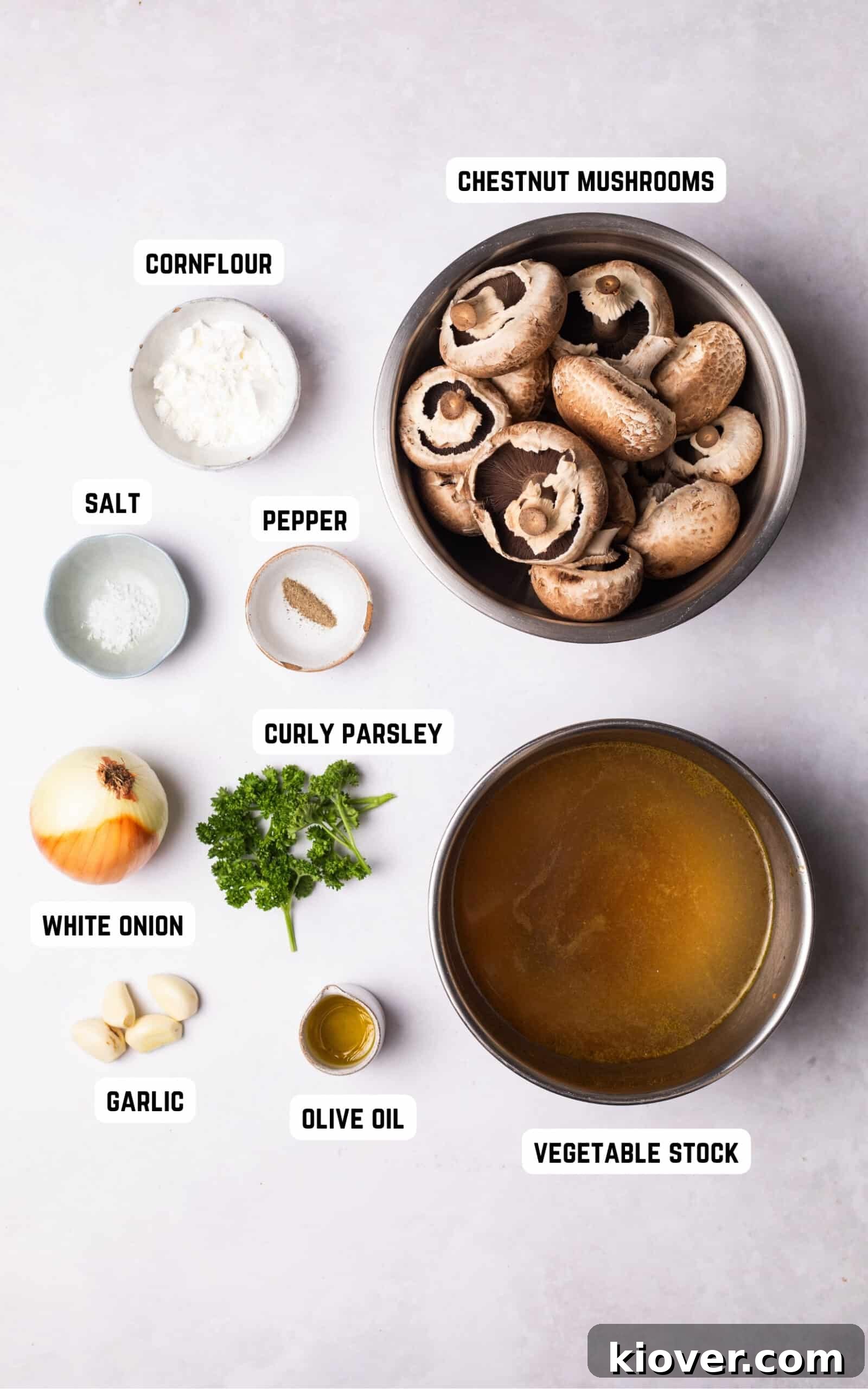 An array of fresh ingredients for mushroom soup, including chestnut mushrooms, white onion, garlic cloves, and curly parsley, neatly arranged on a wooden board.
