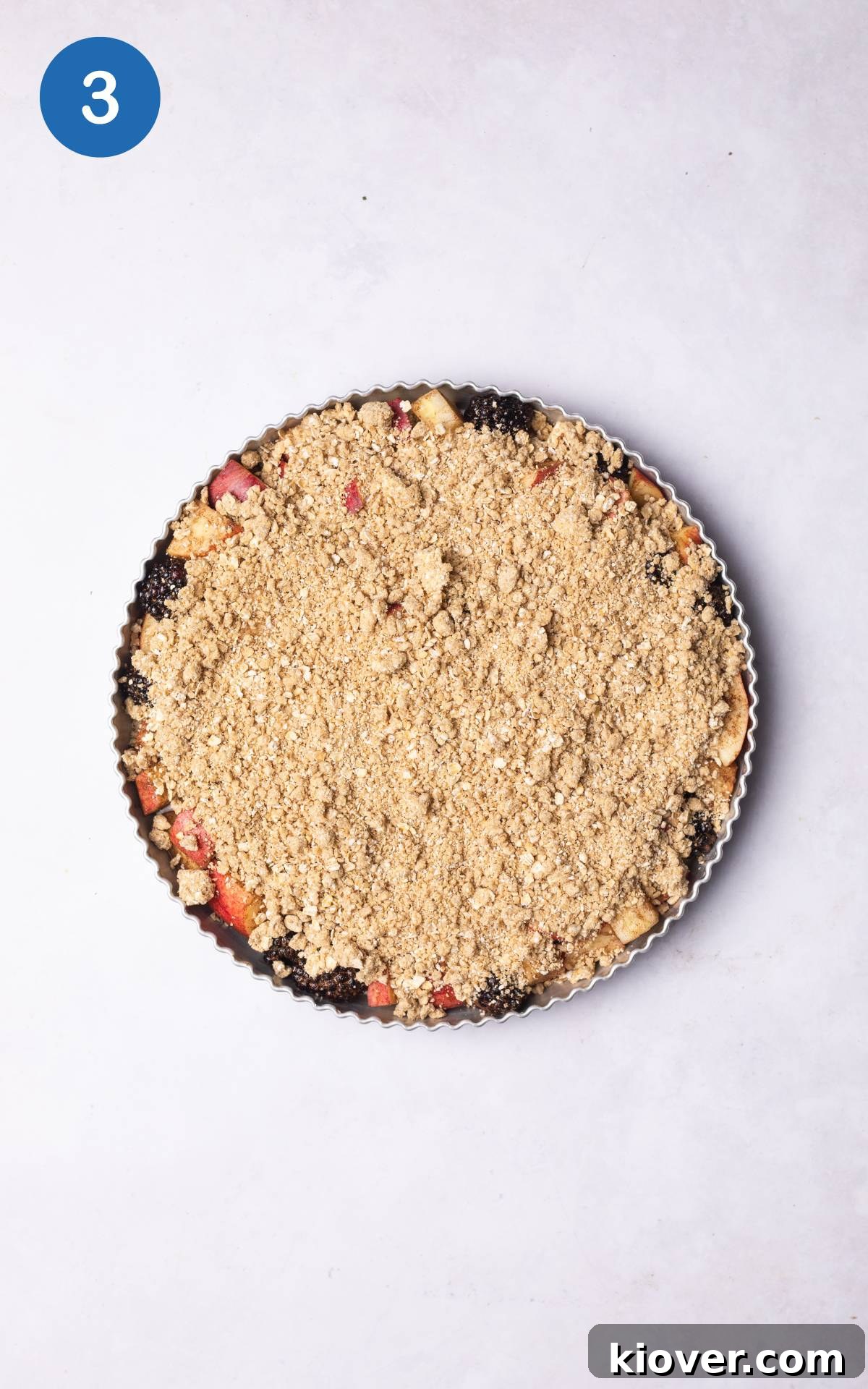 An ovenproof dish filled with the apple and blackberry fruit mixture, generously topped with the prepared oat crumble, ready for baking.