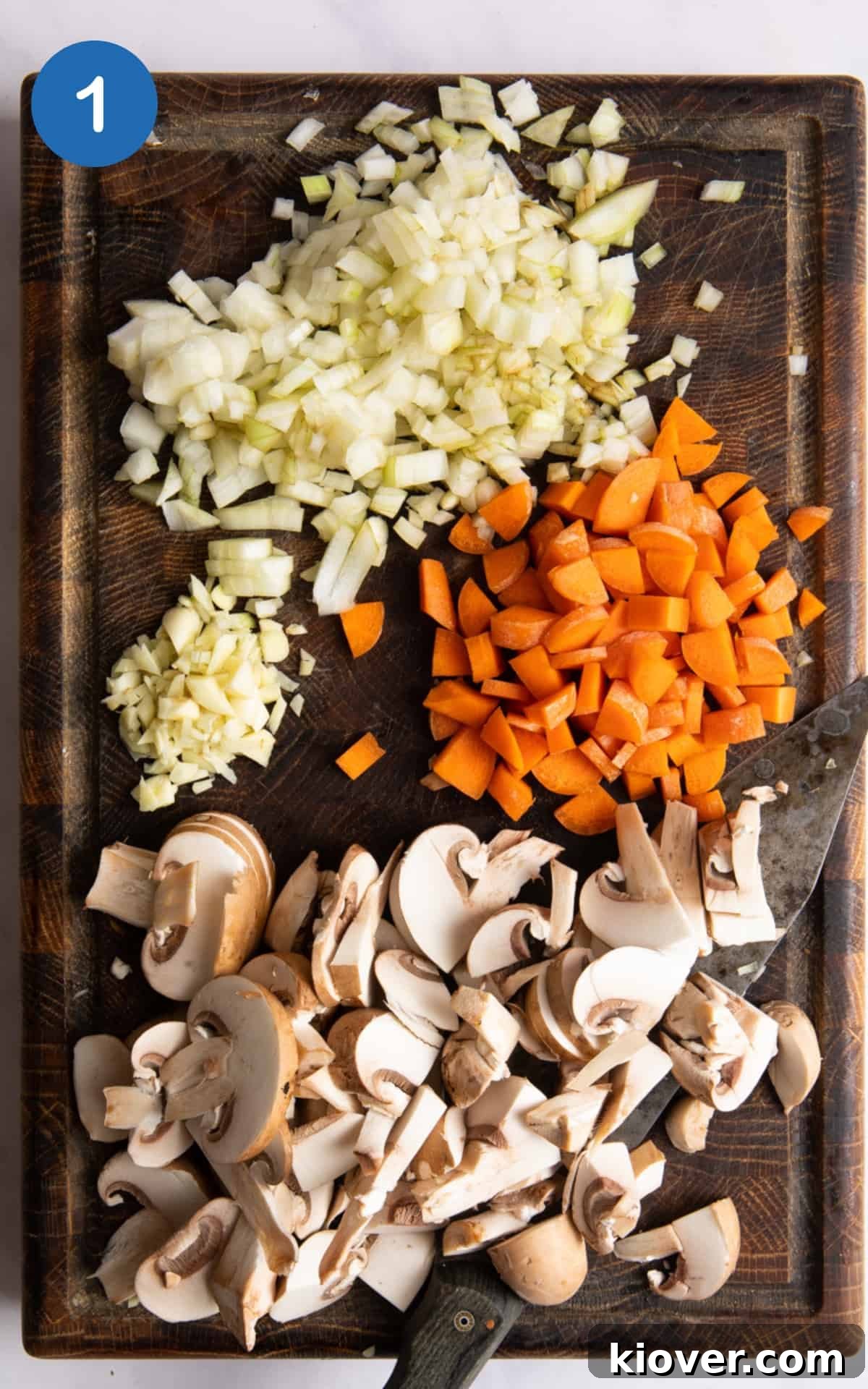 Finely diced onion, crushed garlic, sliced mushrooms, and chopped carrot displayed on a dark wooden chopping board, ready for cooking.