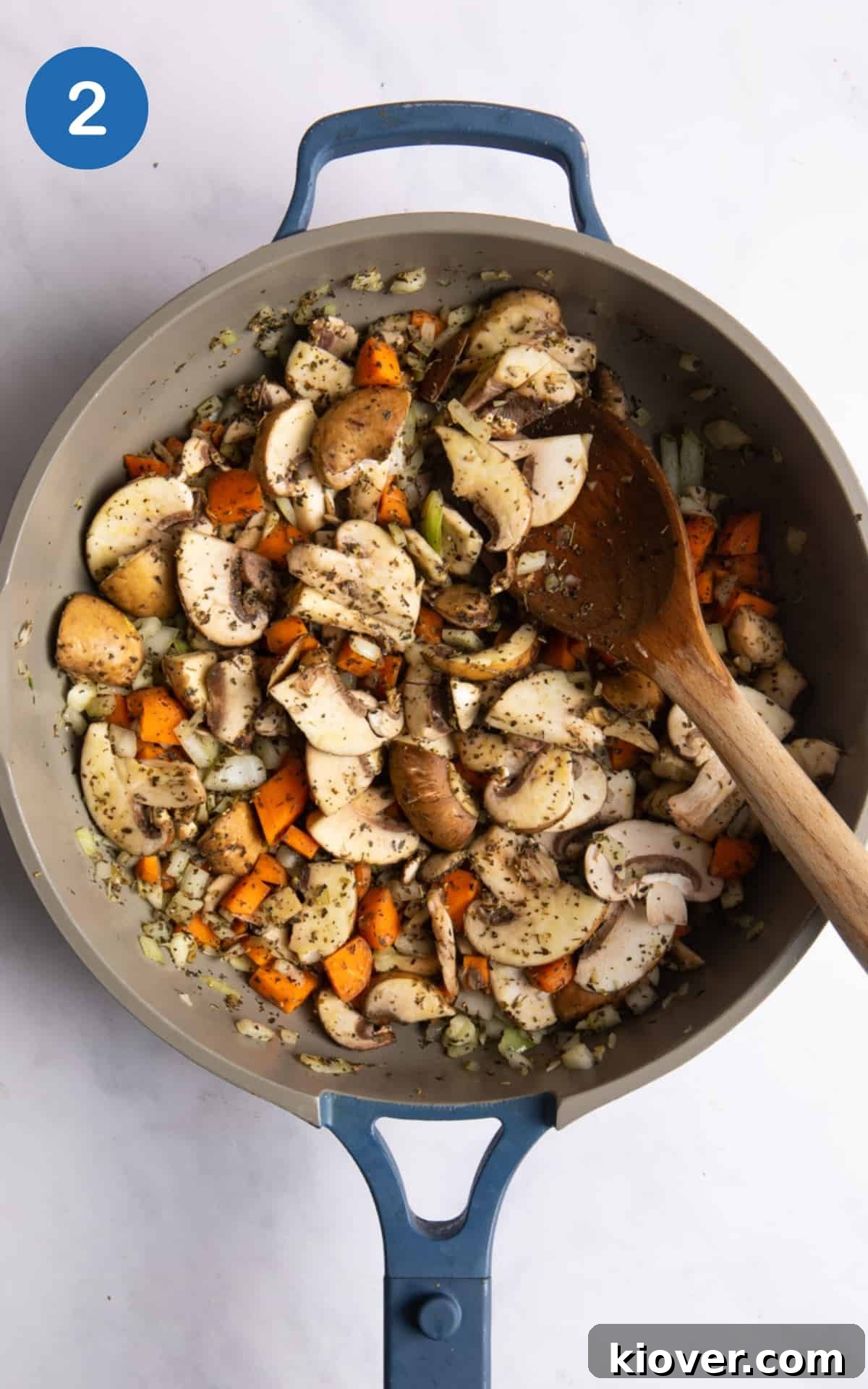 Onions, mushrooms, carrot, and garlic are being sautéed with dried herbs in a large frying pan on a white marble background.