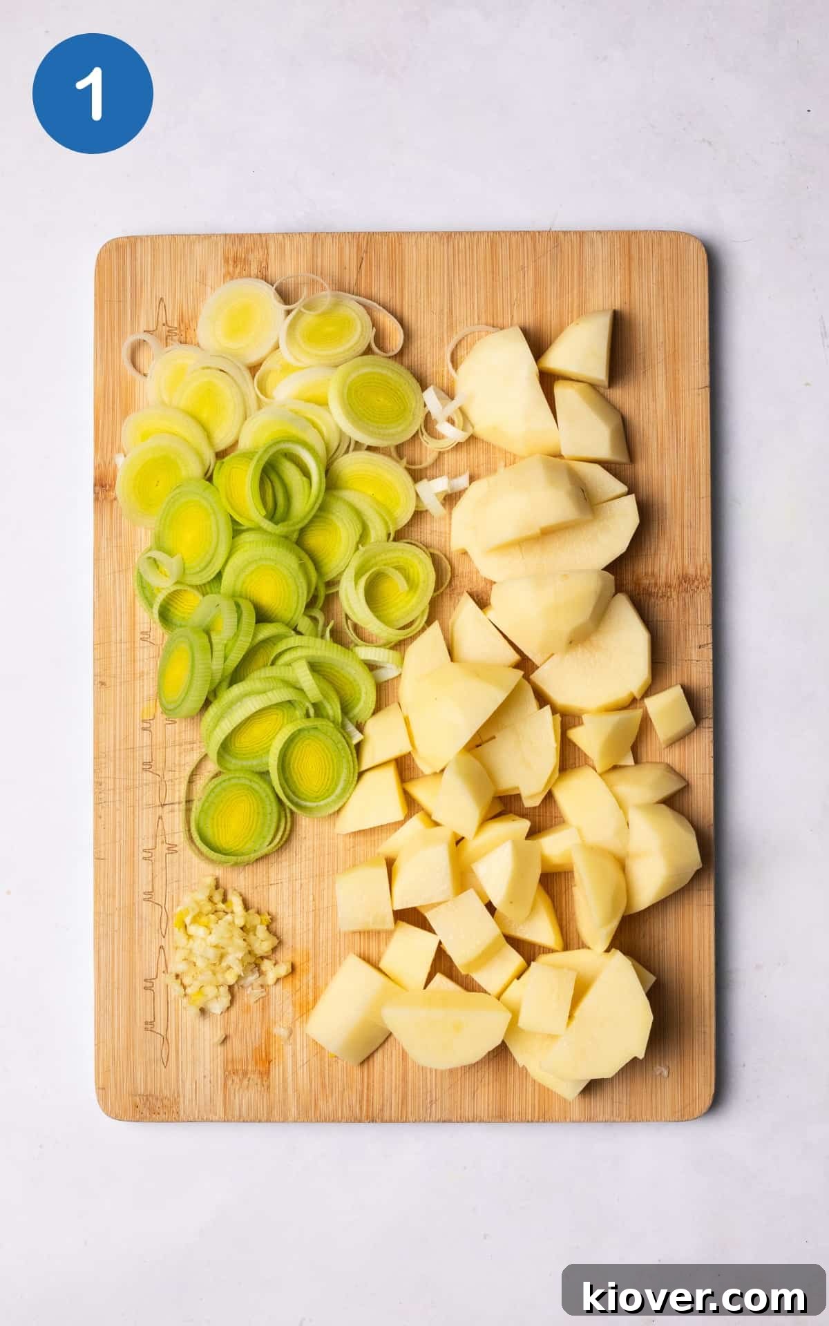 Prepared vegetables: peeled and quartered potatoes, sliced leek, and crushed garlic cloves on a wooden cutting board.