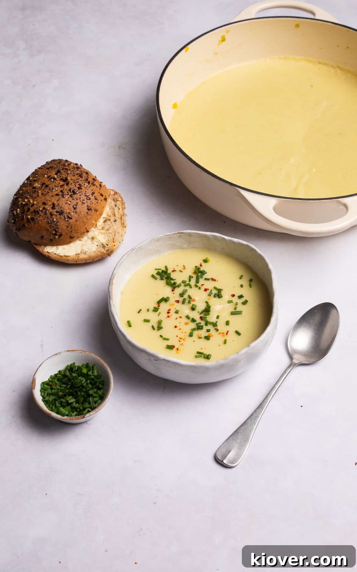 Creamy vegan potato and leek soup served in a bowl, garnished with fresh chives and chili flakes, with a bread bun on the side.