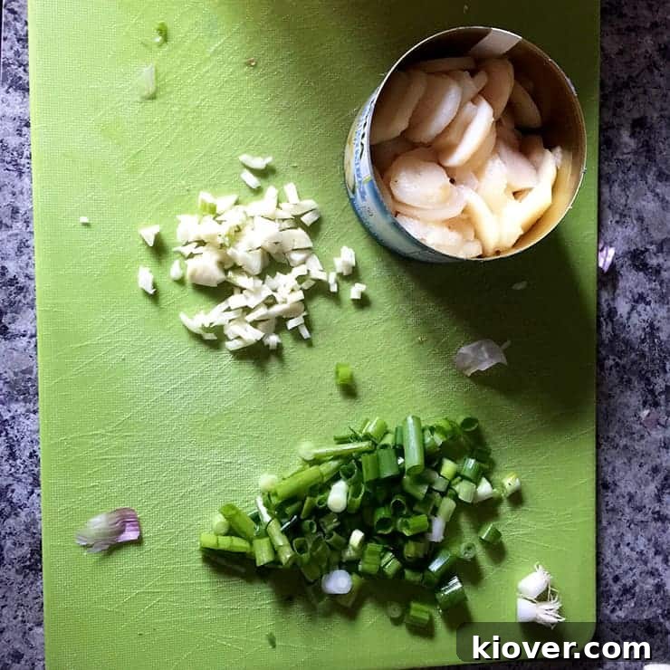 A vibrant selection of water chestnuts, freshly chopped green onions, and minced garlic cloves are artfully arranged on a wooden cutting board, ready for stir-frying.