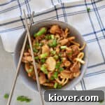 A top-down view of a bowl of crispy chicken chow mein, garnished with green onions. Chopsticks rest elegantly on the side of the bowl, which sits on a plaid napkin, evoking a cozy, authentic dining experience.