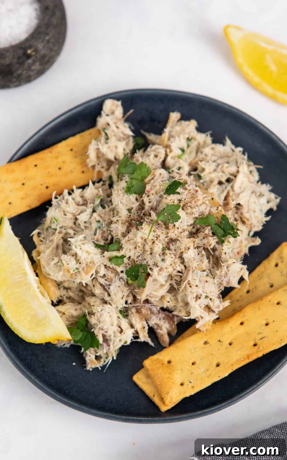A serving of smoked mackerel pate on a round plate, garnished with fresh parsley and a lemon wedge, accompanied by a scattering of crackers.