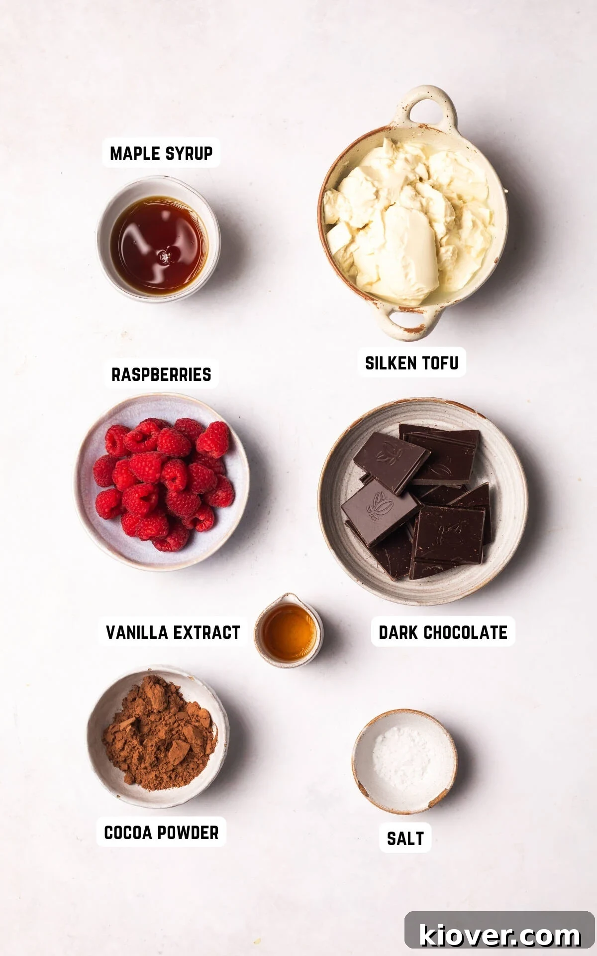 A flat lay photograph showcasing all the fresh ingredients needed to make the Vegan Chocolate Mousse: dark chocolate, silken tofu, vanilla extract, maple syrup, cocoa powder, salt, and fresh raspberries.