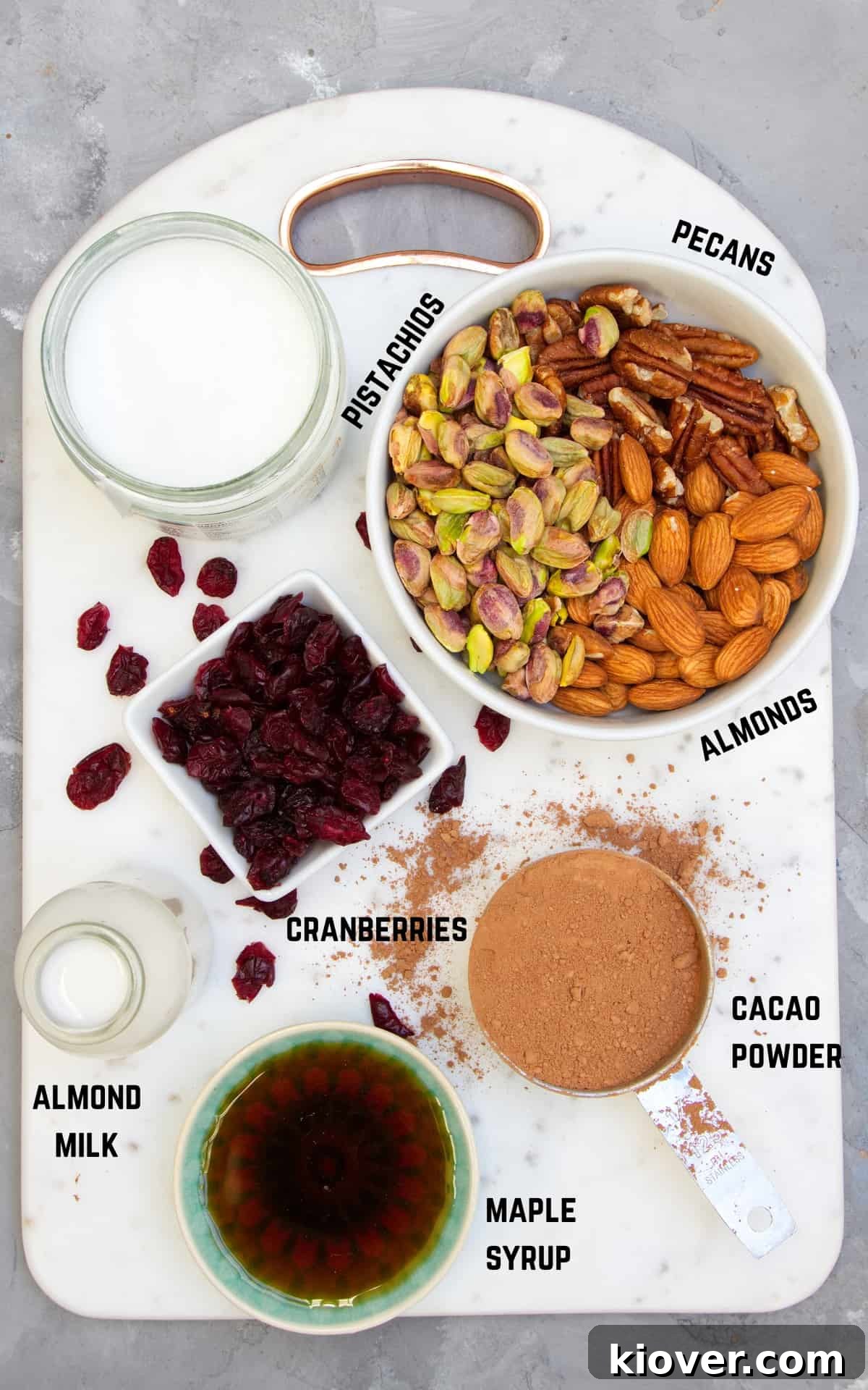 Plant-Based Rocky Road Delights 3 Bowls of cacao powder, maple syrup, coconut oil, almond milk, nuts, and cranberries – ingredients for vegan rocky road – arranged on a white cutting board.