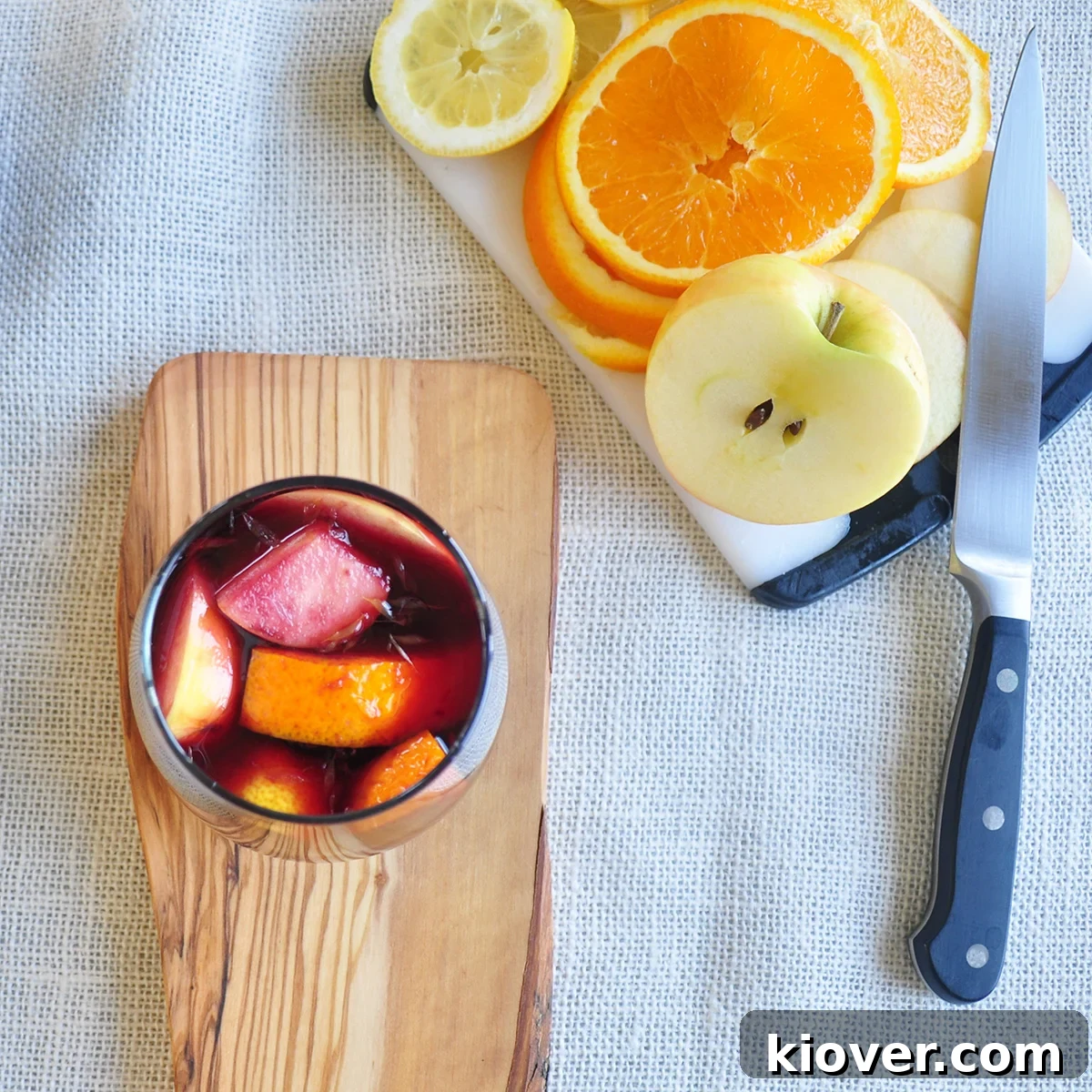 Easy Red Sangria Glass with sangria and fruit in it on a piece of wood. There is a cutting board with oranges, lemons and an apple on it.
