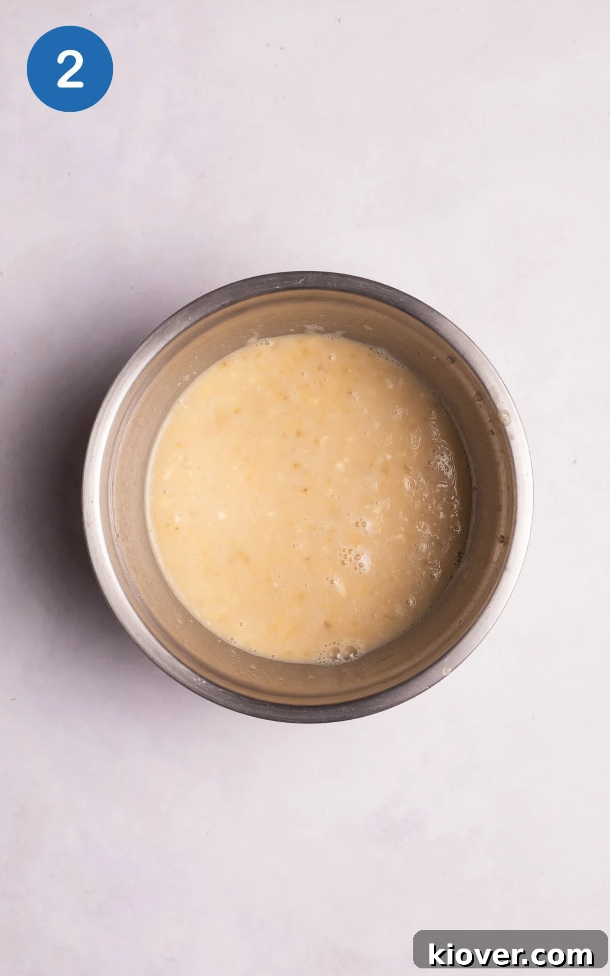 Mashed banana being combined with vanilla extract, maple syrup, and milk in a mixing bowl.