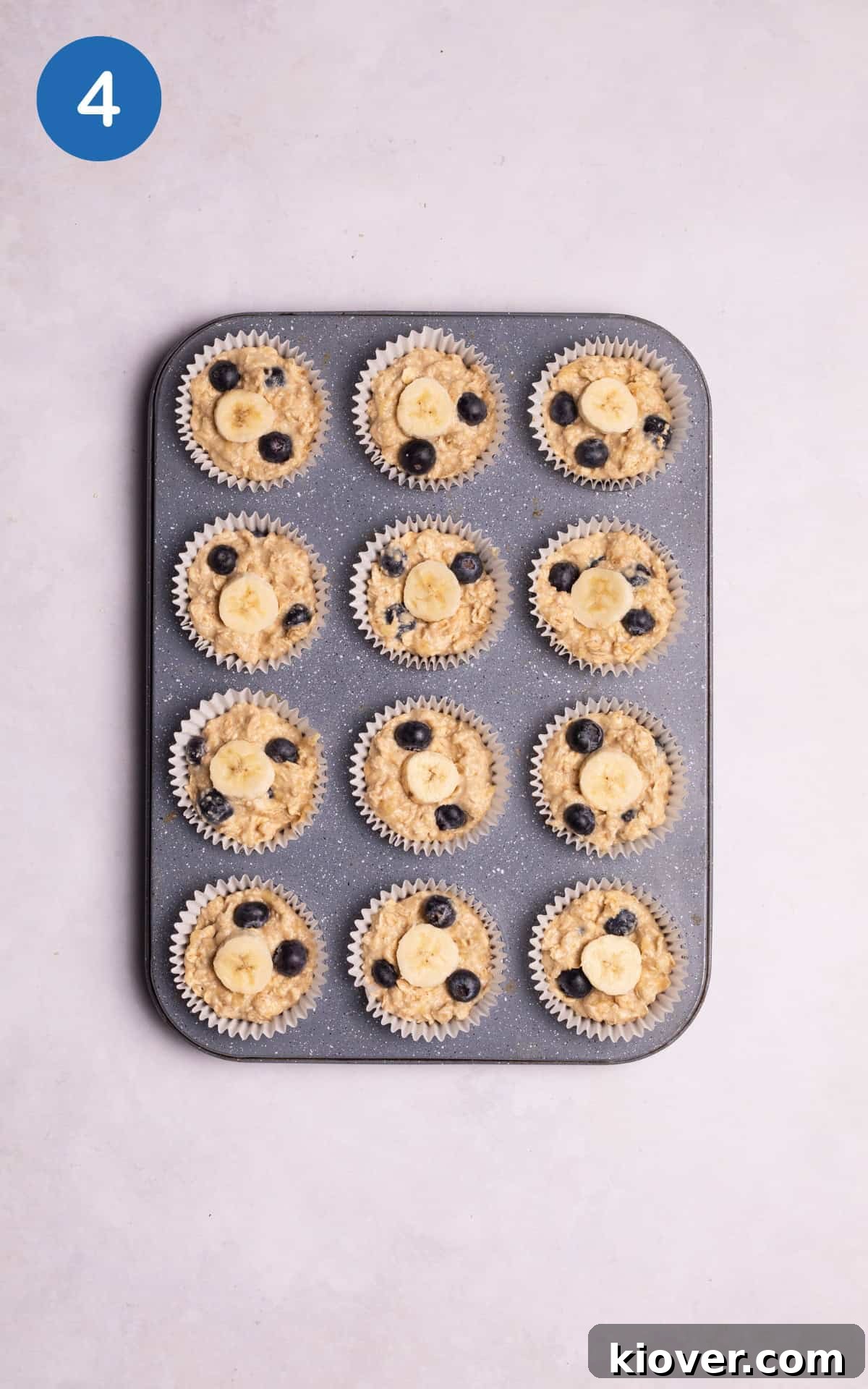 Banana oatmeal muffin mixture scooped into a muffin tin with blueberries and banana slices on top.