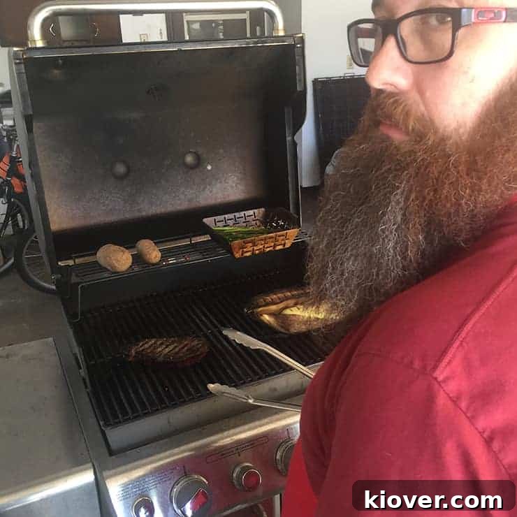 A person tending to a ribeye steak on a searing hot grill, looking back at the camera. The grill is set with different heat zones, and potatoes are also visible on the cooler side.