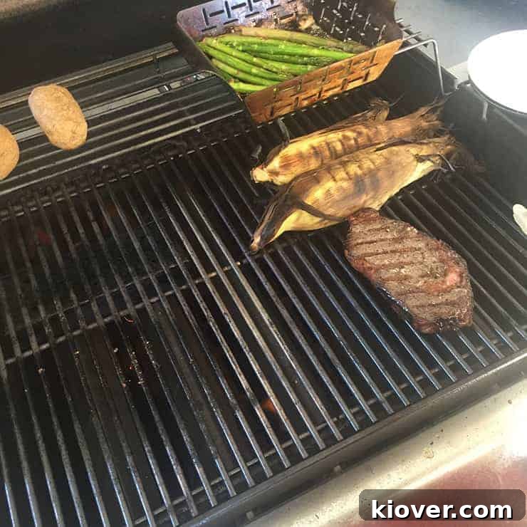 A perfectly grilled ribeye steak resting on the cooler zone of a gas grill, alongside cooking asparagus, corn on the cob, and baked potatoes, showcasing effective zone grilling.
