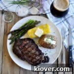 Overhead view of a beautifully grilled ribeye steak on a plate with traditional sides like corn, baked potato, and asparagus.