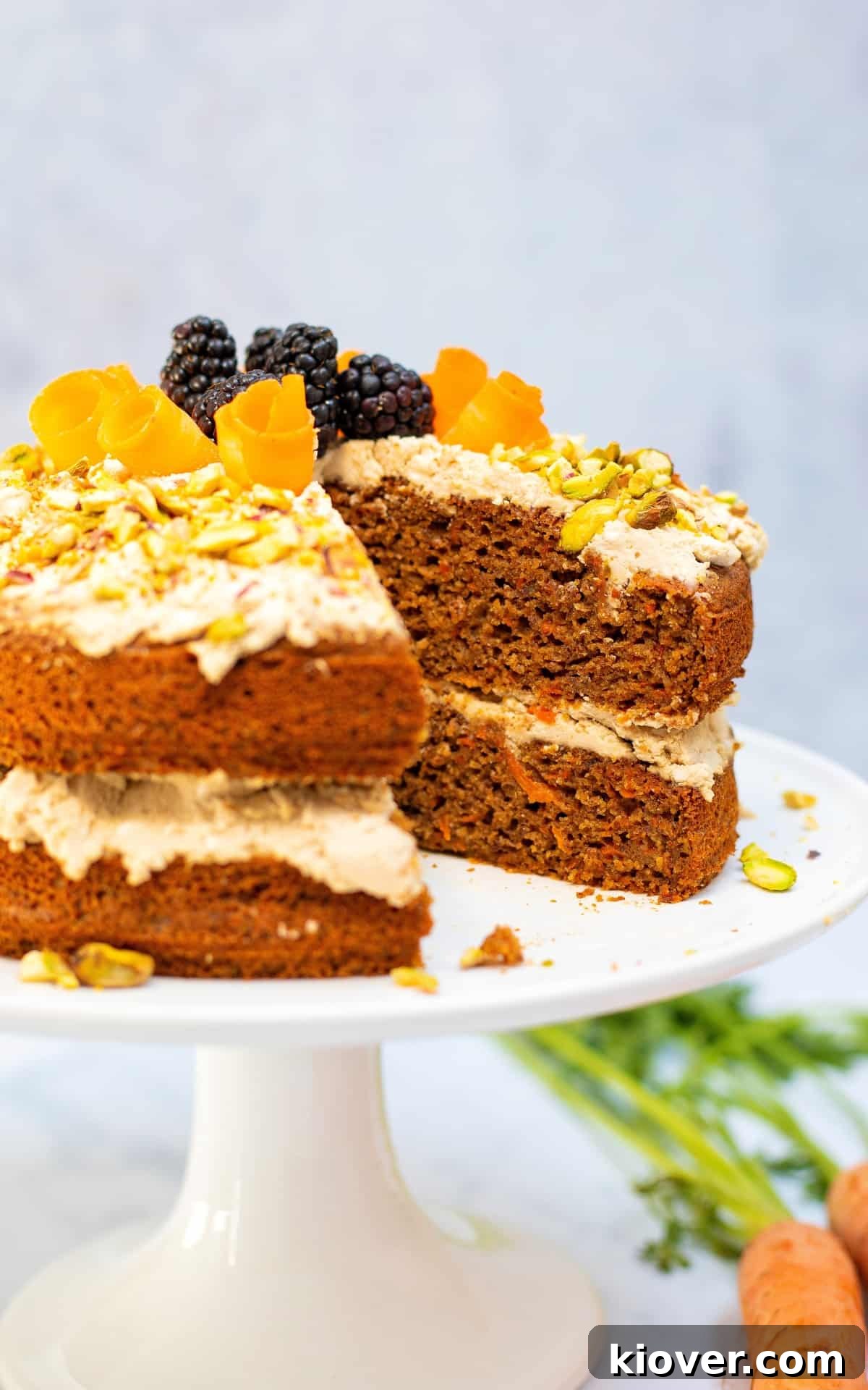 Decadent Gluten-Free Carrot Cake 7 A beautifully decorated two-tiered carrot cake with coconut icing and fresh fruit, with a slice removed, resting on a white cake stand.
