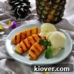 Grilled pineapple with a side of basil ice cream on a plate with pineapple in the background and some decorative flowers.
