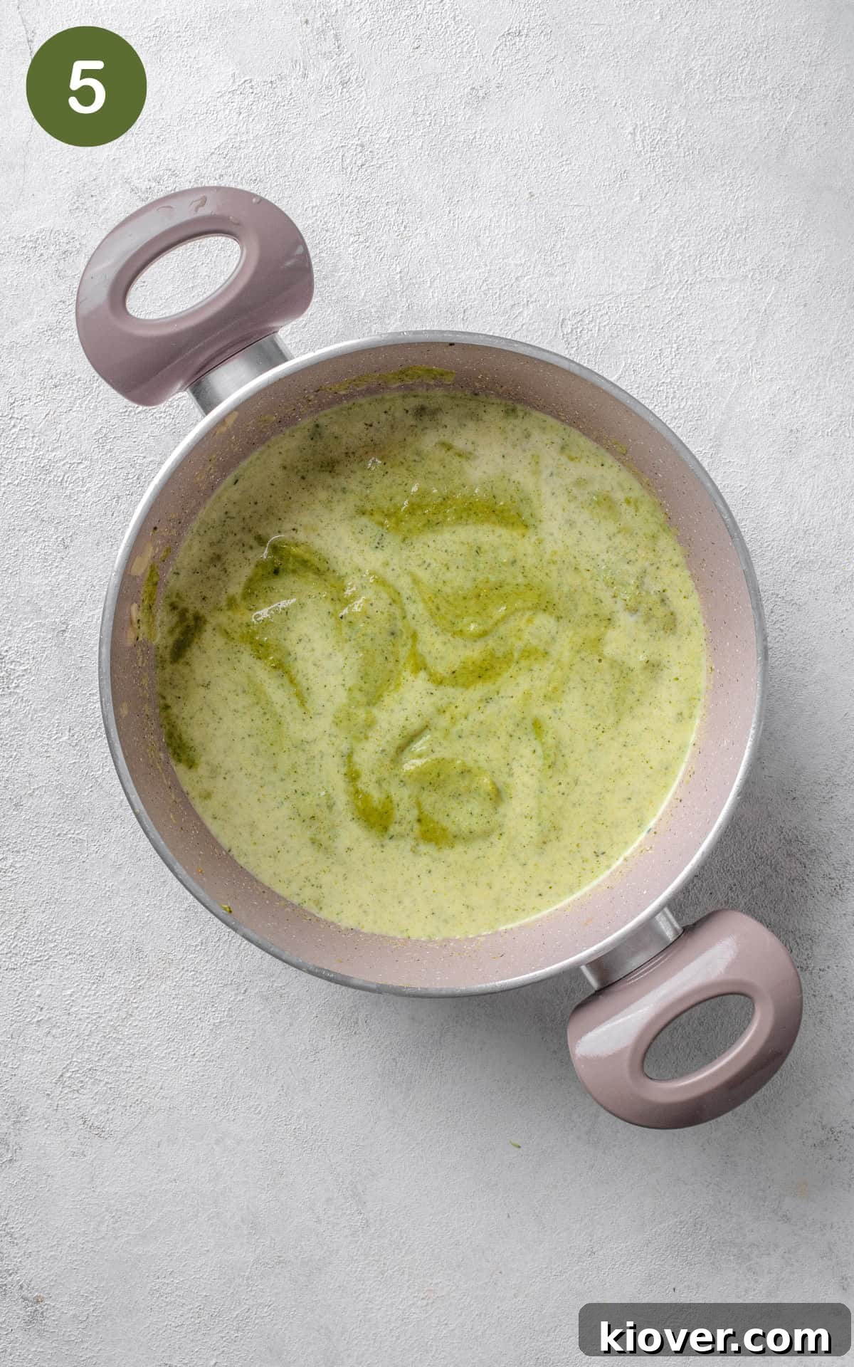 Smooth green broccoli soup in a pot, with a carton of cooking cream being poured in to finish.