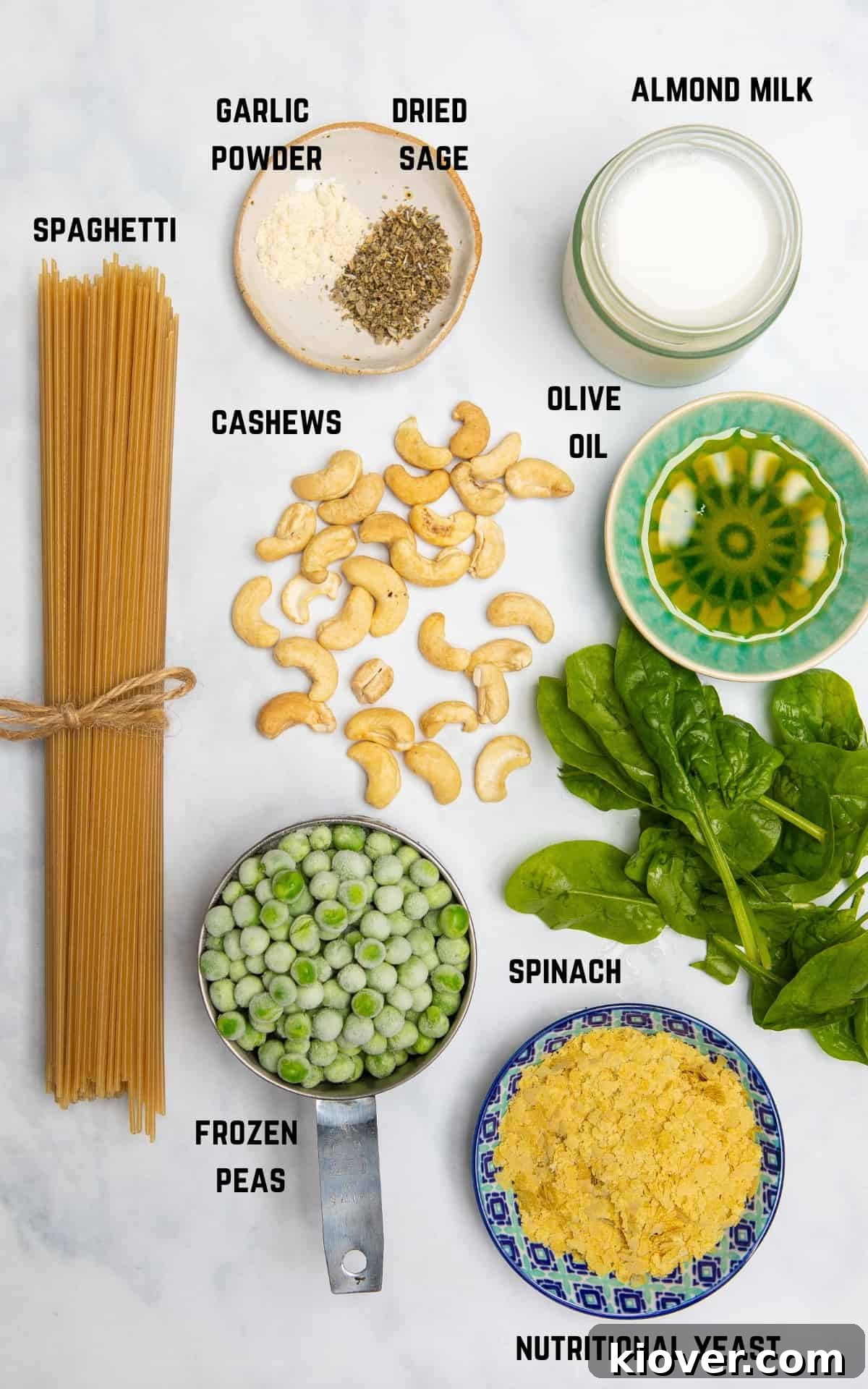 Ingredients to make vegan carbonara laid out on a white surface, including spaghetti, peas, spinach, cashews, nutritional yeast, olive oil, and plant-based milk.