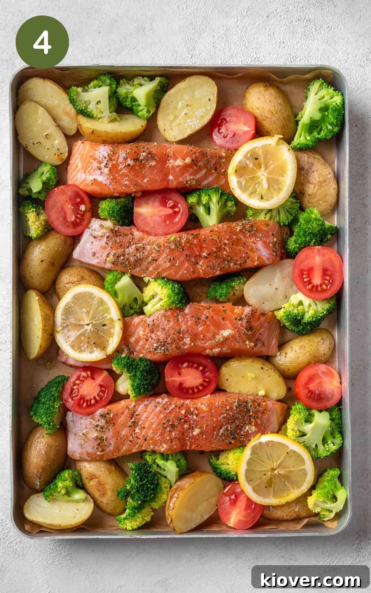 A baking tray filled with roasted baby potatoes, marinated salmon fillets, fresh broccoli florets, and cherry tomatoes, garnished with lemon slices, ready for the final bake.