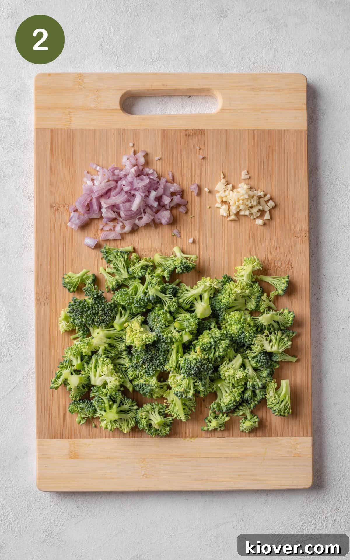 Fresh broccoli florets, chopped shallots, and finely minced garlic on a wooden cutting board, prepped for cooking.