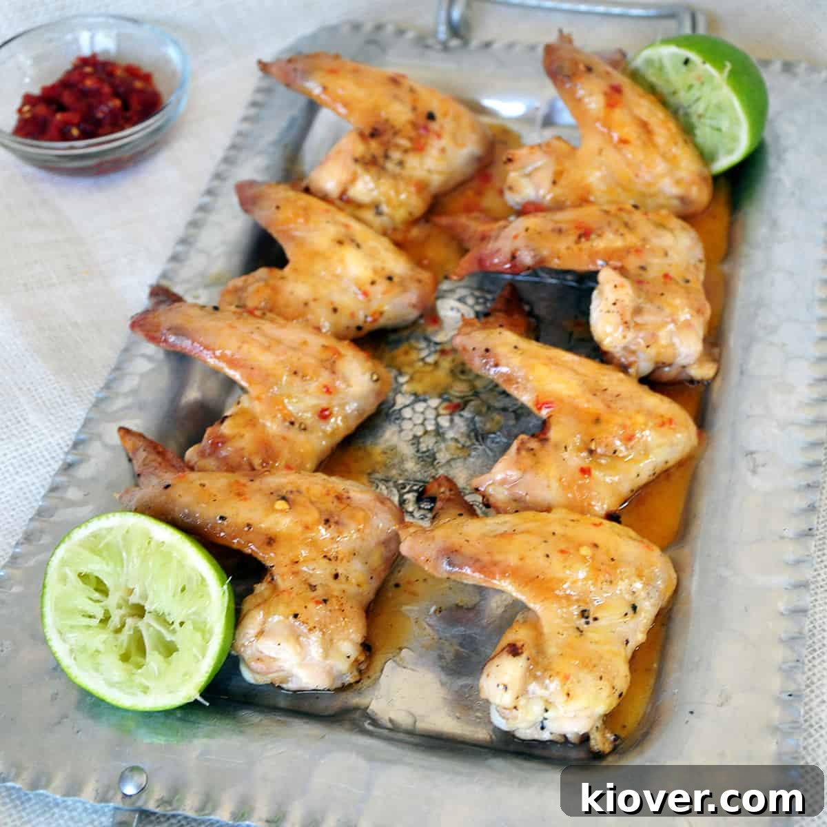 Tequila Lime Chicken Wings Eight perfectly grilled Tequila Lime Chicken Wings arranged on a gleaming silver platter, ready for serving. Accompanying them are two squeezed lime halves and a small bowl of fiery Sambal Oelek in the background, hinting at the delightful flavors.