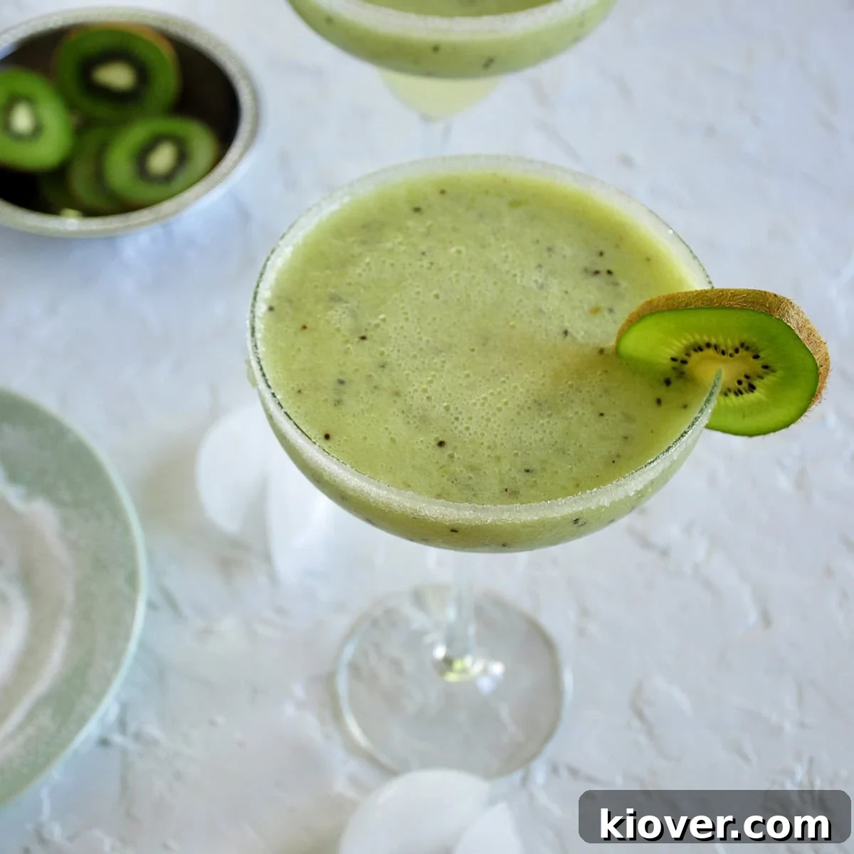 Vibrant Frozen Kiwi Coconut Margarita garnished with a fresh kiwi slice, presented on a wooden board with ice, more sliced kiwi, and a plate of sugar for rimming.