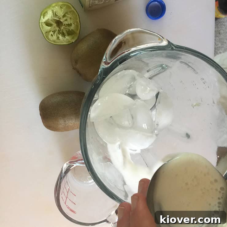 Pouring creamy coconut milk into a blender filled with ice, preparing for a tropical frozen drink.