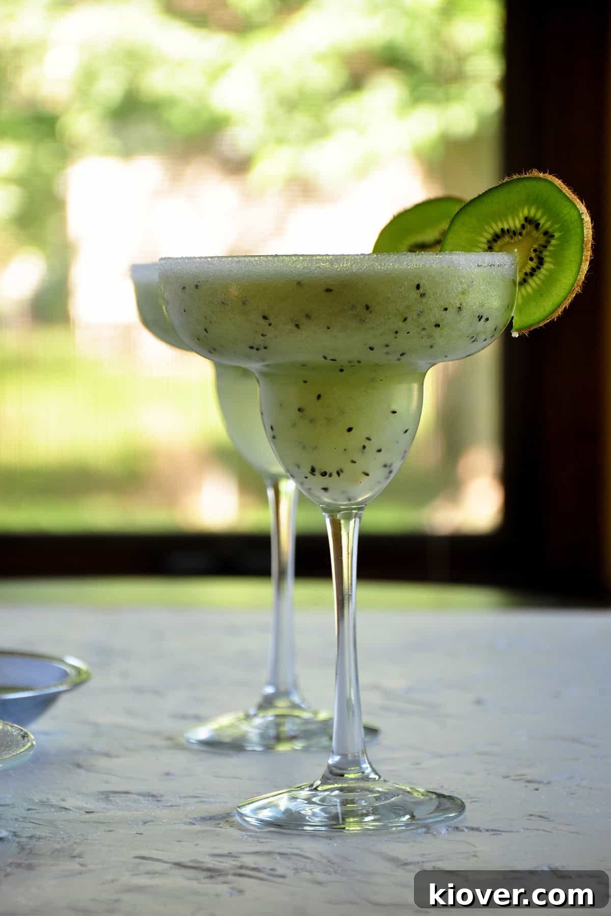 Two perfectly blended Frozen Kiwi Coconut Margaritas, elegantly presented in tall glasses with kiwi slices as garnish, set against a bright window backdrop, inviting a refreshing sip.