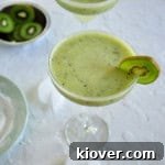 Frozen Kiwi Coconut Margarita with a kiwi slice as garnish. There is some ice laying on the board below along with a dish of sliced kiwi and a plate of sugar that was used for rimming the glass