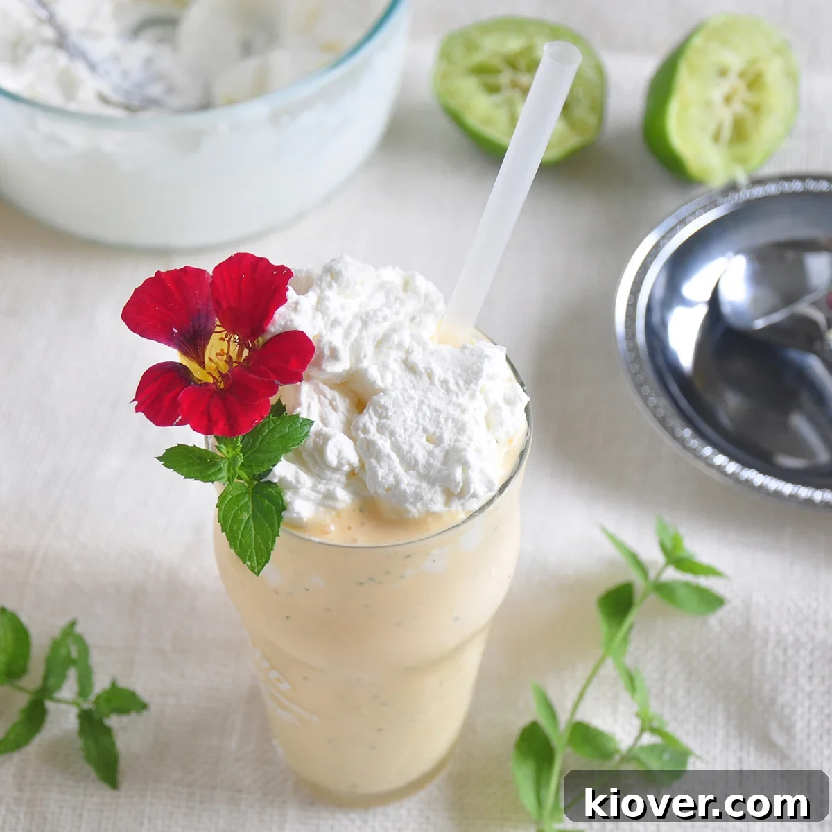 The Mojitarita Milkshake - a mint margarita dessert on a white cloth with fresh mint leaves and lime wedges scattered around the glass. The drink is a vibrant green, topped with whipped cream and garnished with a mint sprig, viewed from a high angle.