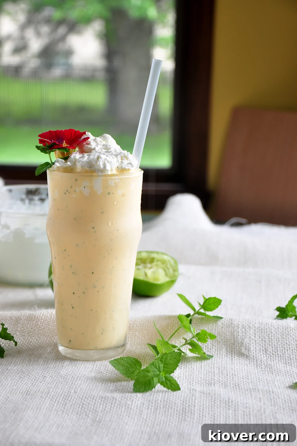 This an Ice Cream Mint Margarita and shot from the side. There is mint and lemon in the background, as well as a window. The Mojitarita milkshake is in a tall glass, garnished with fresh mint and a lime wedge, sitting on a light surface with a window and plants in the soft-focus background.