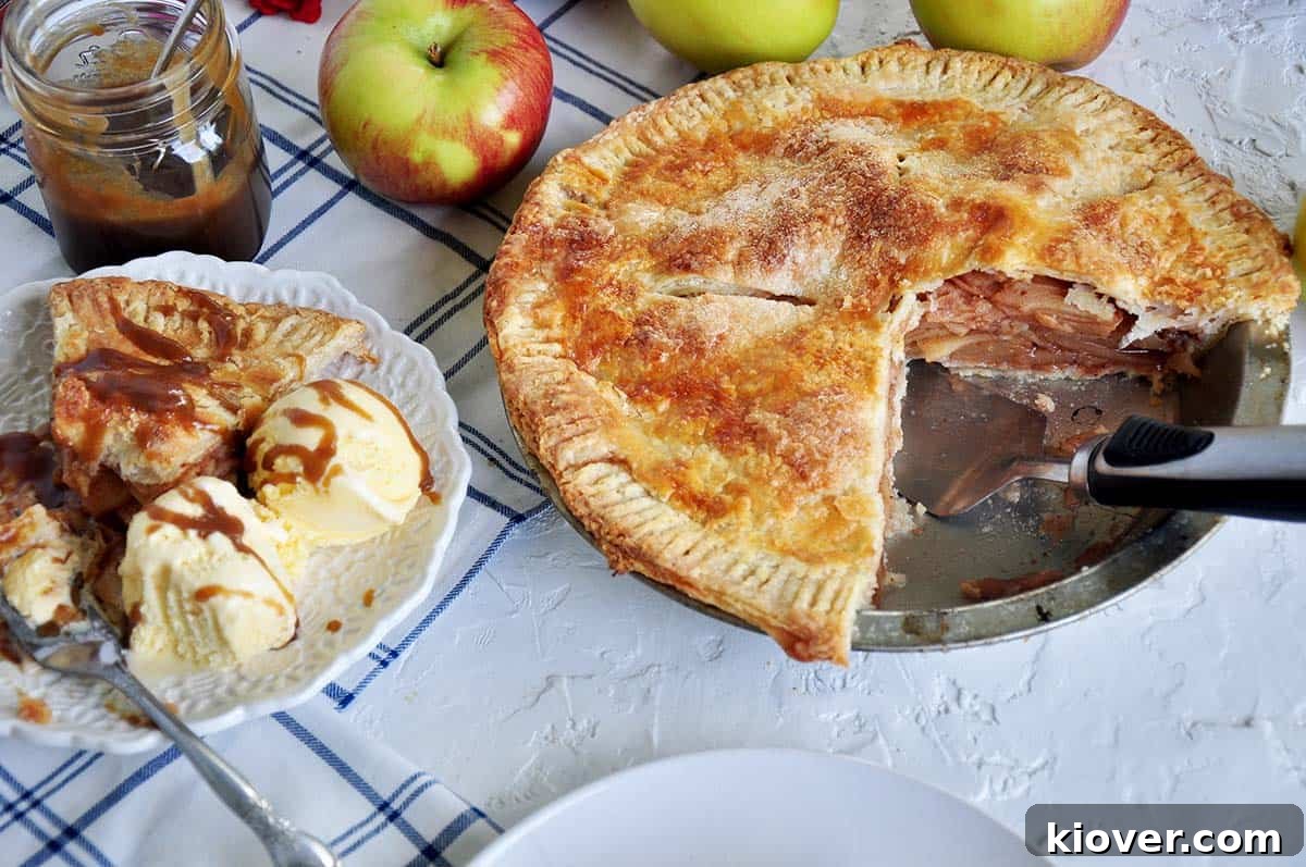 Baked apple pie with two pieces removed and placed on a plate. There is a serving utensil in the pie pan. Several Braeburn apples in the background