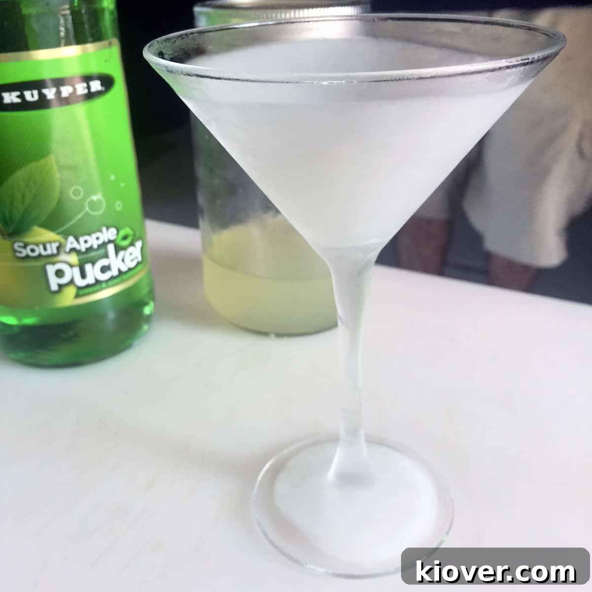 A frozen martini glass, glistening with frost, with a bottle of Sour Apple Pucker liqueur visible in the softly blurred background.