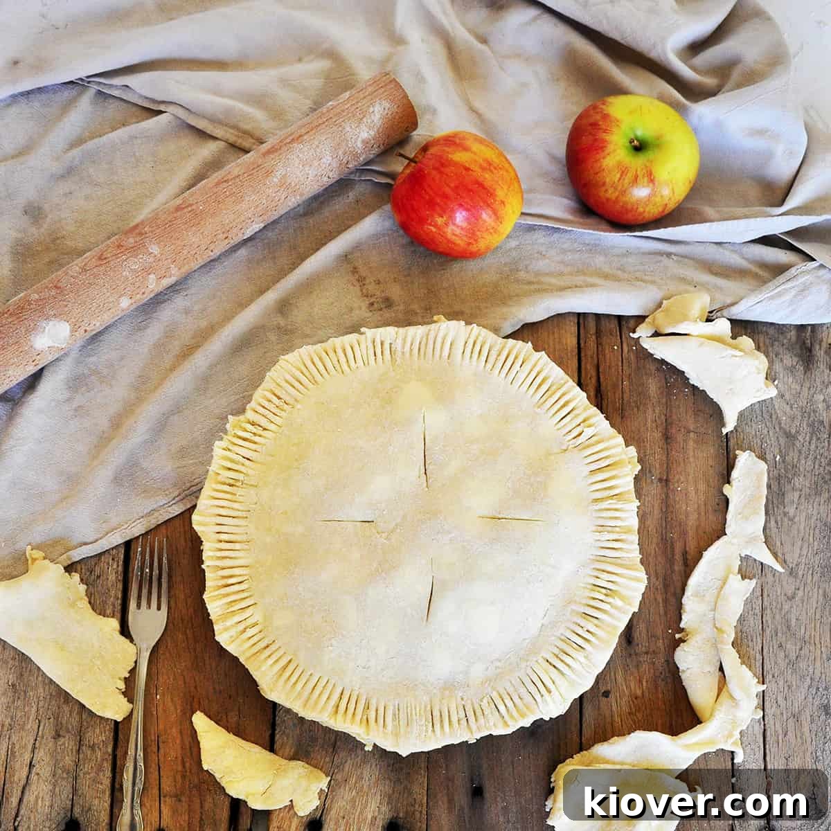 A Raw Pie Crust with apples in the pie. It is on a picnic table with two Braeburn apples, a rolling pin, and is laying on a cloth.