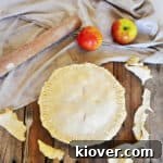 A Raw Pie Crust with apples in the pie. It is on a picnic table with two Braeburn apples, a rolling pin, and is laying on a cloth.