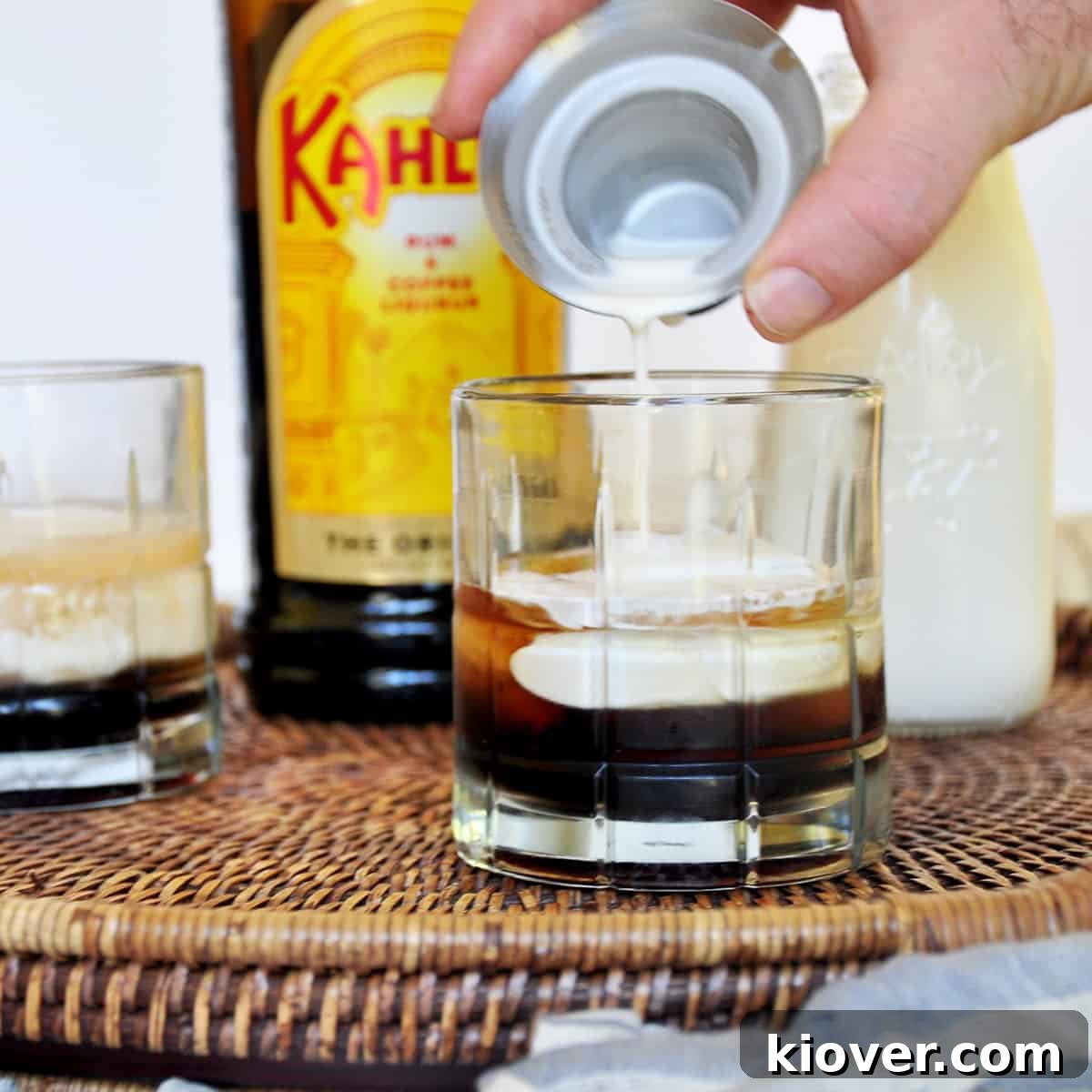 Delicately pouring rich, white heavy cream over the back of a spoon into a glass filled with vodka and coffee liqueur, creating a beautiful layered effect in a White Russian cocktail.