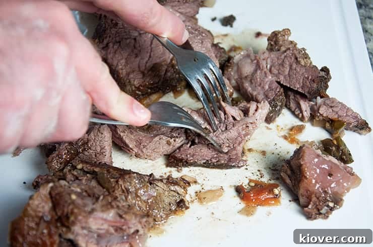 Two forks expertly pulling apart a large piece of tender, slow-cooked beef on a wooden cutting board, showcasing its easily shreddable texture.