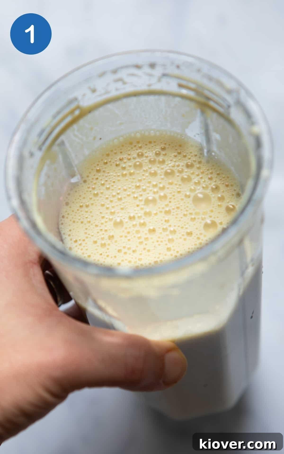A hand holding a blender cup filled with perfectly blended, creamy ingredients for the vegan cheese sauce, set against a pristine white background.