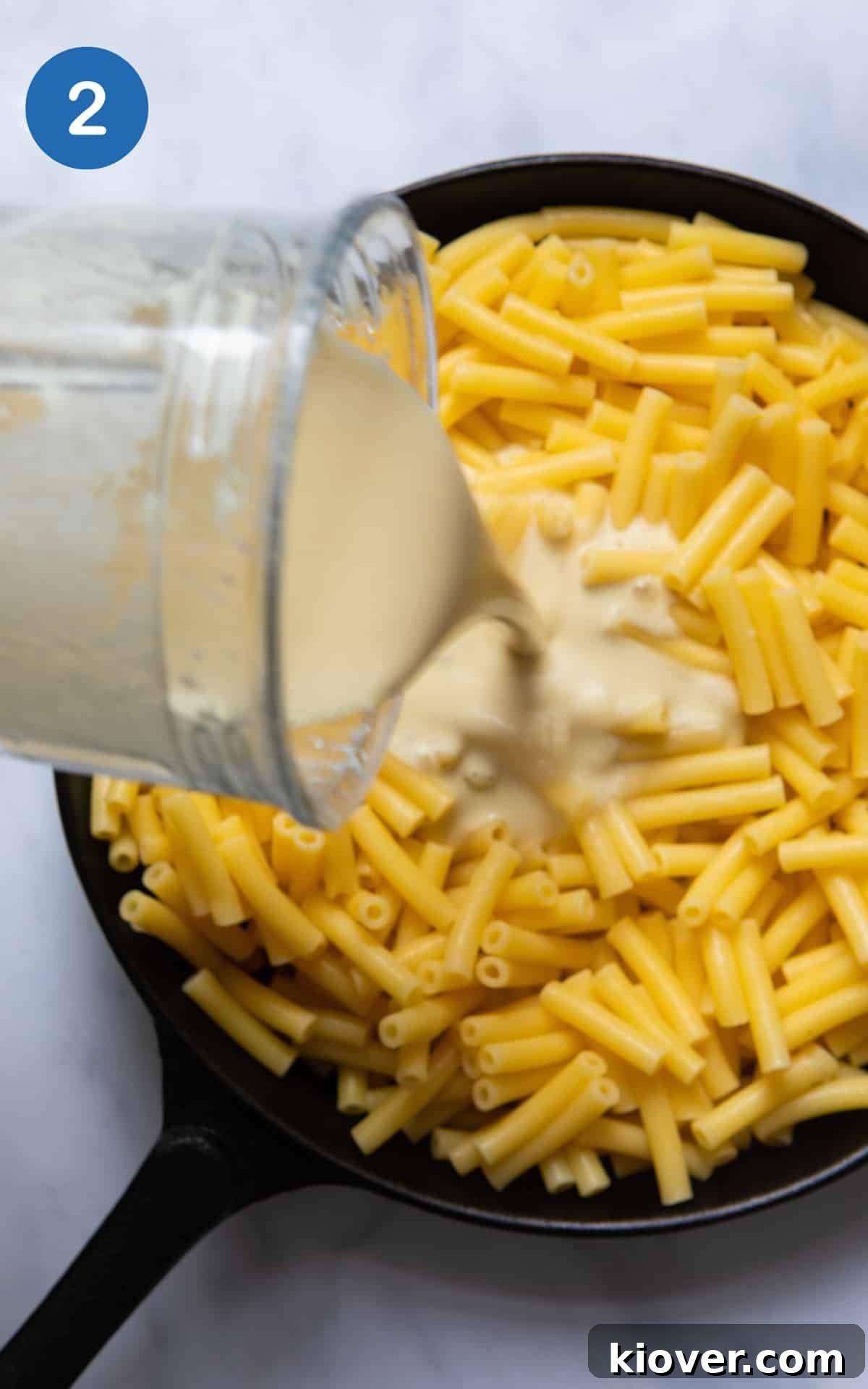 Freshly drained macaroni pasta resting in a black skillet pan, with the creamy vegan cheese sauce being elegantly poured over it, ready for mixing.