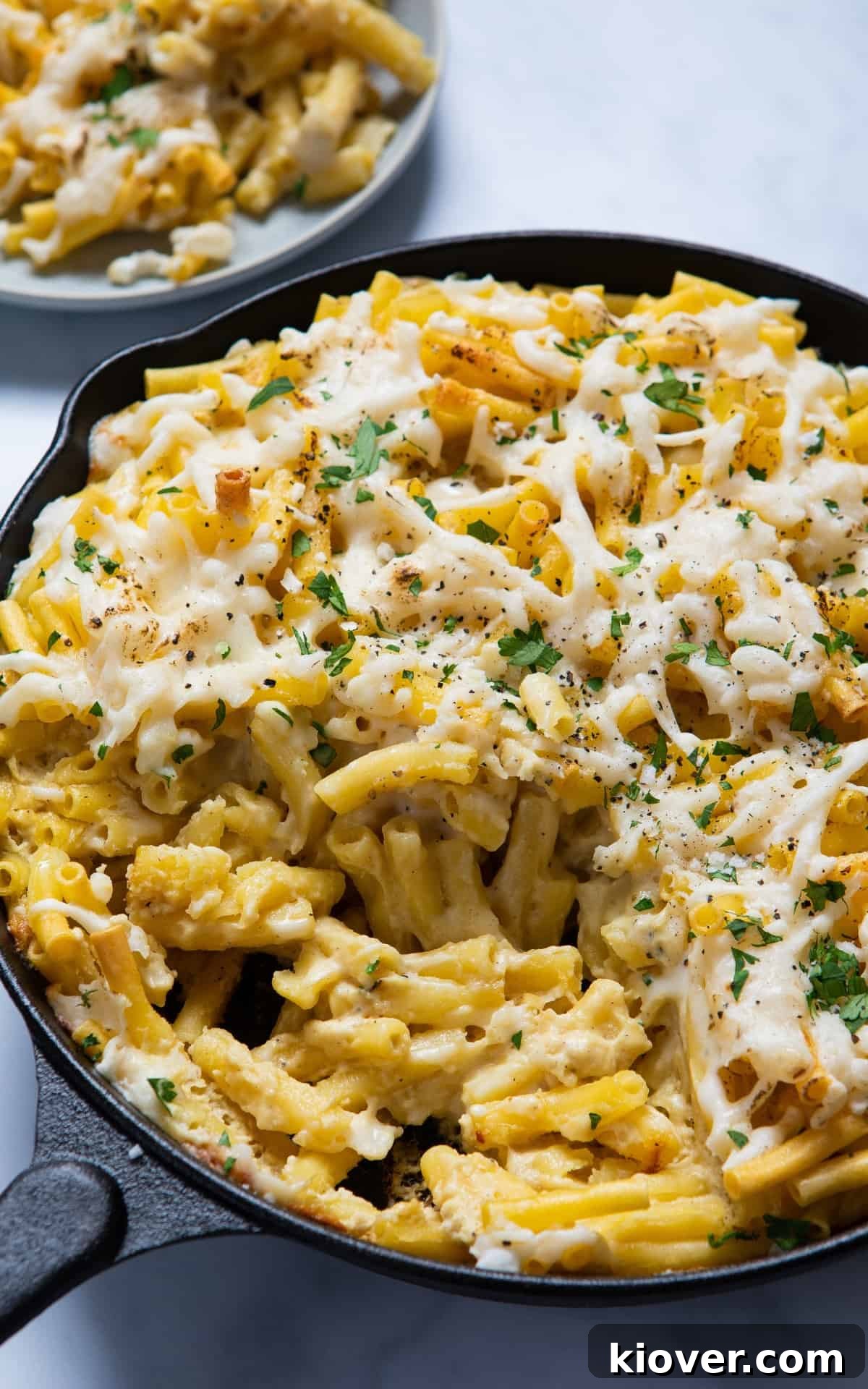 A close-up shot of rich, golden vegan mac and cheese in a black skillet pan, with a plated serving visible in the softly blurred background, all on a crisp white surface.