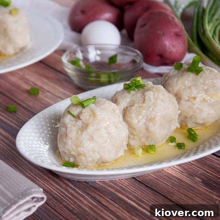 Three perfectly formed Klubb dumplings, with ingredients like shredded potatoes and a block of ham blurred in the background, suggesting the preparation process.